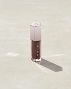 A closed tube of Gloss Bomb Universal Lip Luminizer in the shade Hot Chocolit on a grey background.