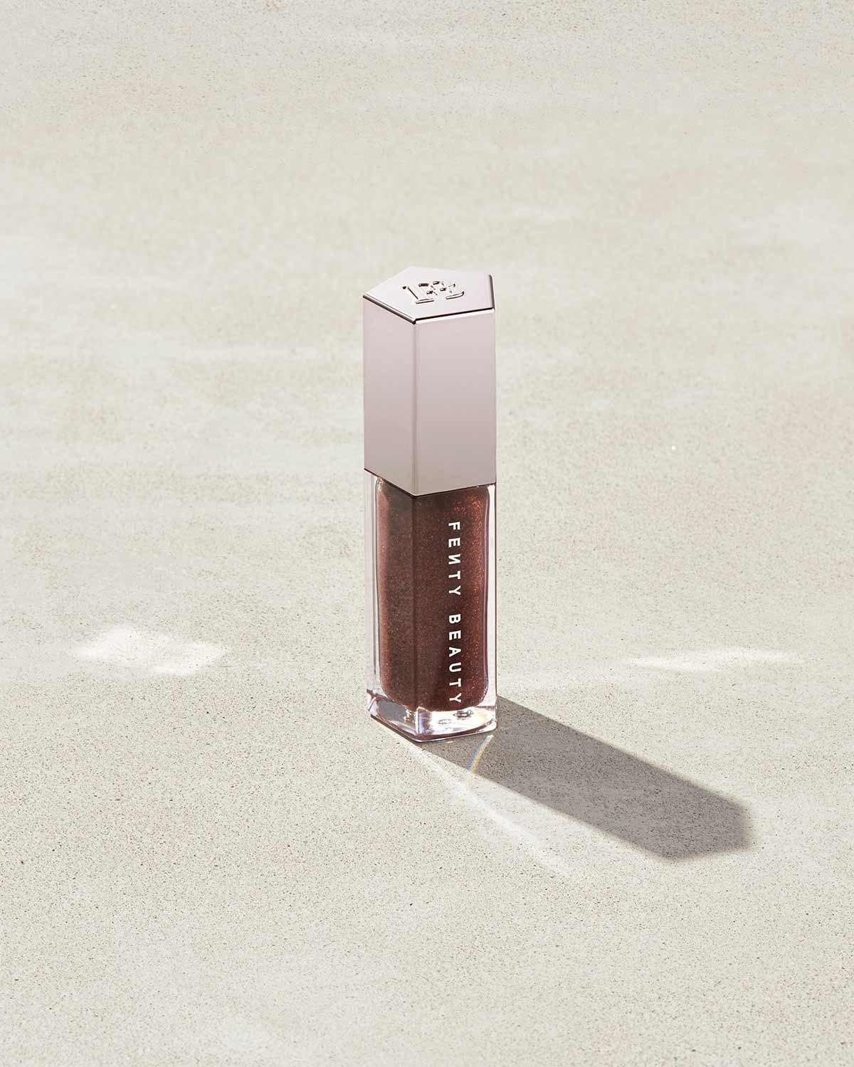 A closed tube of Gloss Bomb Universal Lip Luminizer in the shade Hot Chocolit on a grey background.
